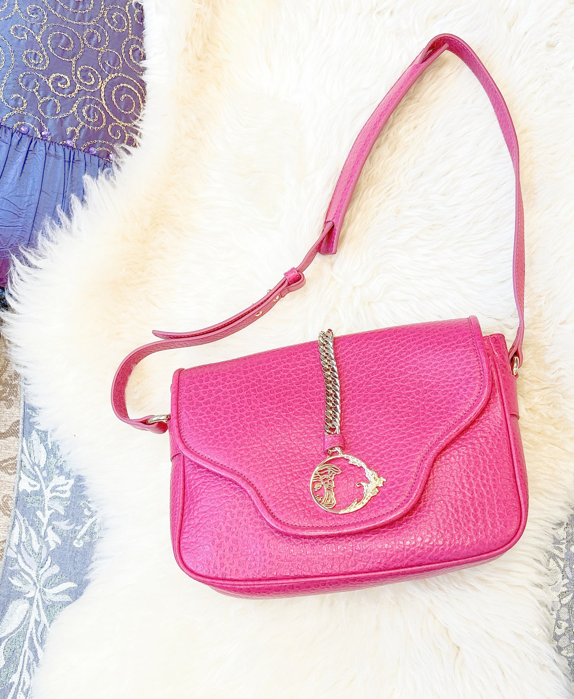 Versace leather bag in vibrant pink with adjustable strap and decorative clasp, placed on a soft white background.