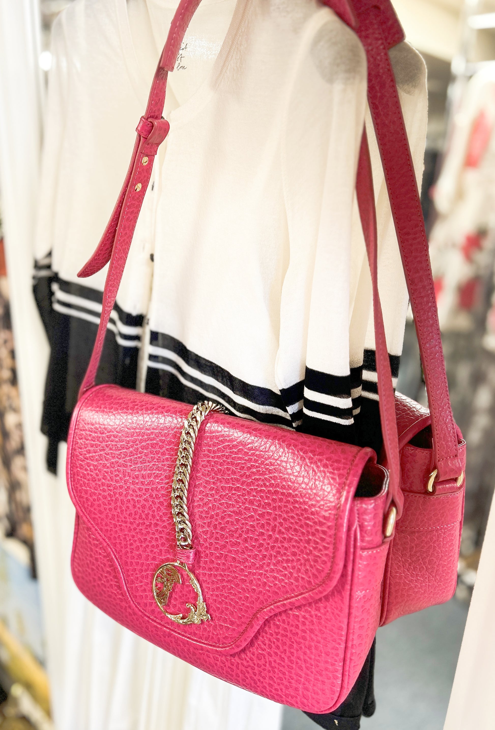 Stylish pink leather handbag with chain detail, hung beside a white and black sweater in a boutique setting.