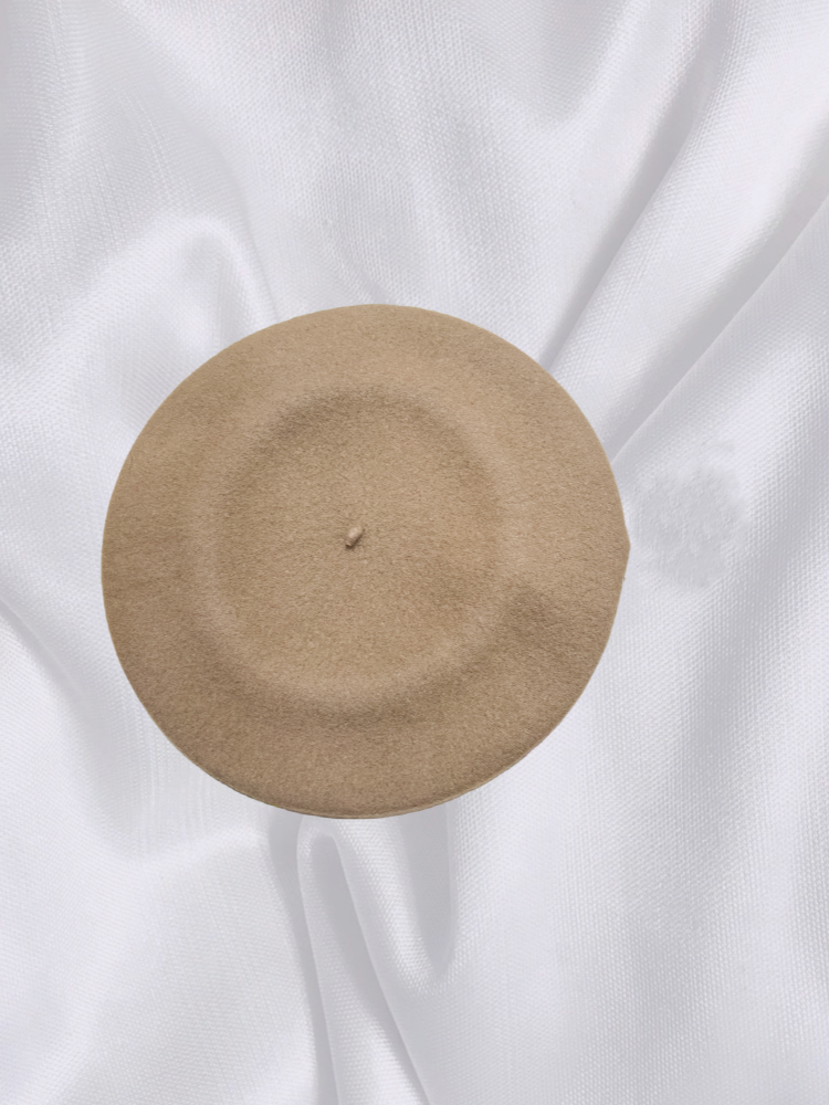 Top view of a beige 100% wool beret on a soft white fabric backdrop, showcasing its smooth texture and round shape.