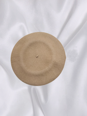 Top view of a beige 100% wool beret on a soft white fabric backdrop, showcasing its smooth texture and round shape.